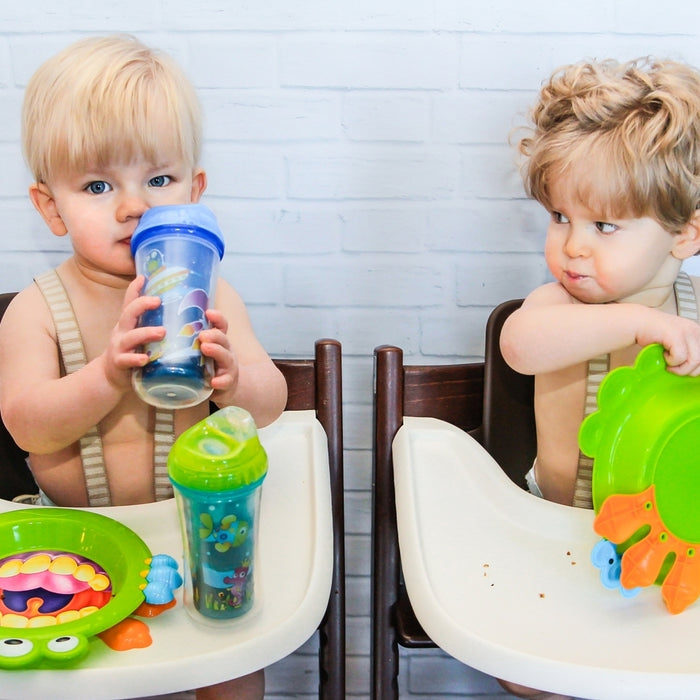 Two toddlers in high chairs are pictured; the child on the left drinks from a blue sippy cup featuring the Clik-it Insulated Cool Sipper Replacement Spout (2 Pack), while colorful dishes and cups sit on their trays against a white brick wall backdrop.