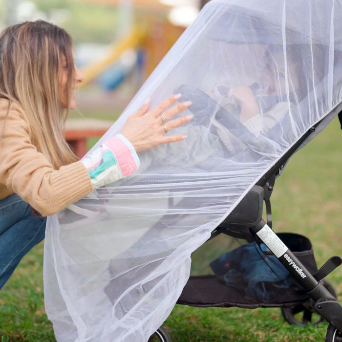 A woman adjusts the Eco Stroller Weather Shield & Insect Netting Set over a baby in a stroller while outdoors in a park.