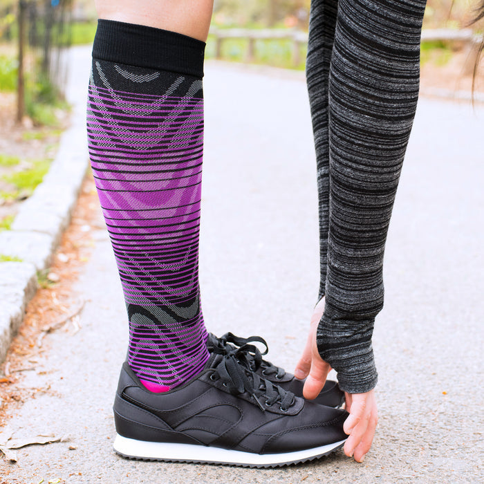 Compression socks with an ombre design in purple and black, worn with black athletic shoes on a pathway.
