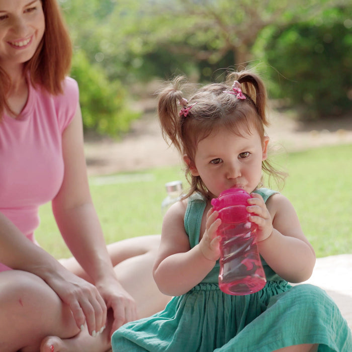 A young girl with pigtails drinks from a Nuby Thirsty Kids Sip It Sport Beginner Travel Cup | Rocket outdoors on a sunny day, sitting next to a smiling woman. Green grass and trees are in the background.