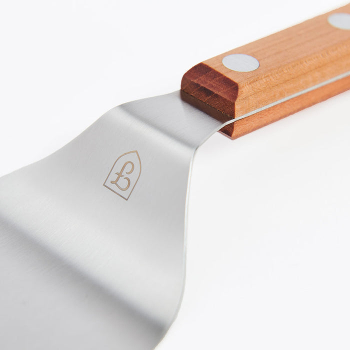 Close-up of the Field Griddle Turner's stainless blade, etched shield logo, and riveted cherry wood handle