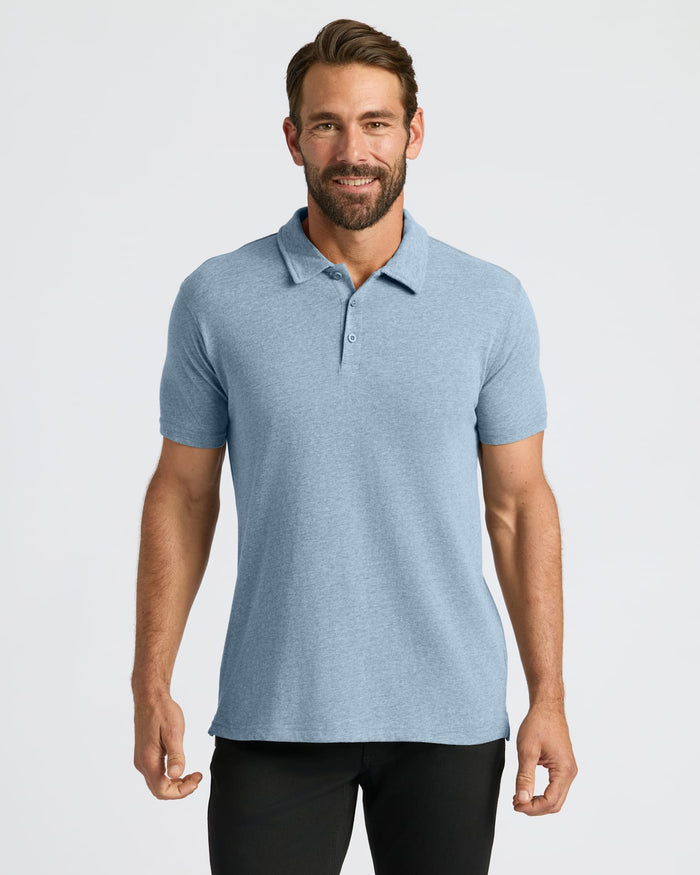 A man wearing a light blue heathered short-sleeve polo shirt, paired with black pants, standing against a solid light gray studio background.