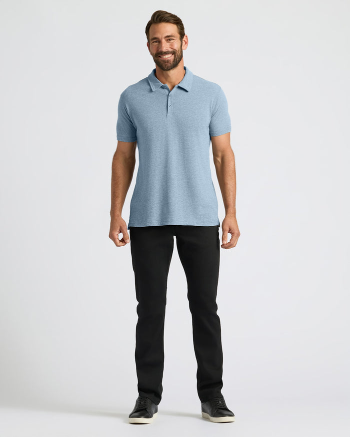 A man modeling a light blue short-sleeve polo shirt with black pants and black sneakers, standing against a plain light gray studio background.