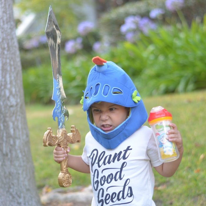 A young child stands on grass in a "Plant Good Seeds" shirt, holding a large toy sword and a sippy cup fitted with the Clik-it Insulated Cool Sipper Replacement Spout (2 Pack), while wearing a blue toy knight helmet.