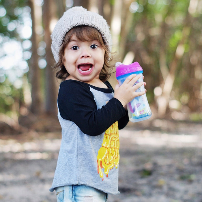 A young child outdoors, trees blurred behind, smiles while holding a colorful sippy cup fitted with the Clik-it Insulated Cool Sipper Replacement Spout (2 Pack), wearing a knit beanie, gray shirt with black sleeves, and denim shorts.