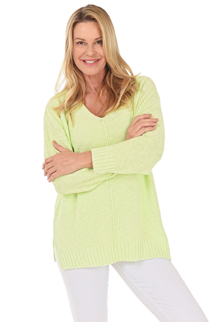 A woman wearing a long sleeve green cotton V-neck sweater, with a seam down the front and white pants.