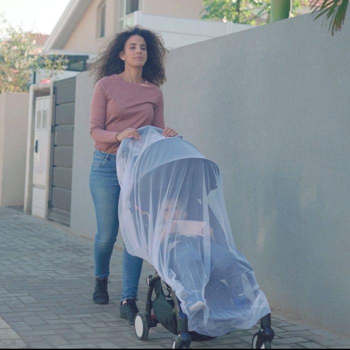 A woman in a pink long-sleeve shirt and jeans pushes the Eco Stroller Weather Shield & Insect Netting Set, crafted from recycled materials, on a sidewalk outside a light-colored house.