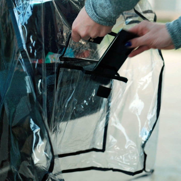 A person places a black smartphone into the Eco Stroller Weather Shield & Insect Netting Set, a transparent, recycled-material bag with a black-edged pocket. The bag is partially open, and they are wearing a gray long-sleeve top.
