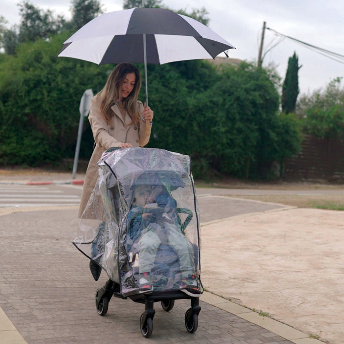 A woman in a beige coat pushes a stroller fitted with the Eco Stroller Weather Shield & Insect Netting Set while holding a black-and-white umbrella on a paved path surrounded by greenery.