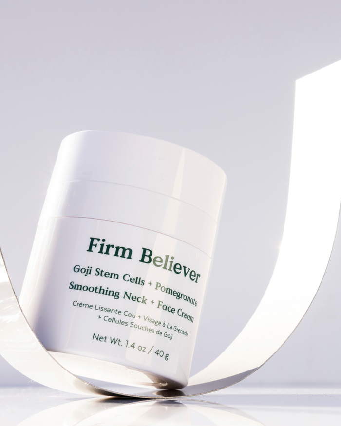 Jar of Firm Believer Goji Stem Cell + Pomegranate Smoothing Neck + Face Cream | Three Ships skincare product placed on a reflective surface with soft natural lighting.