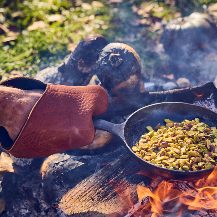 Person in a Field leather oven mitt holds a skillet of pistachios over a smoky open campfire