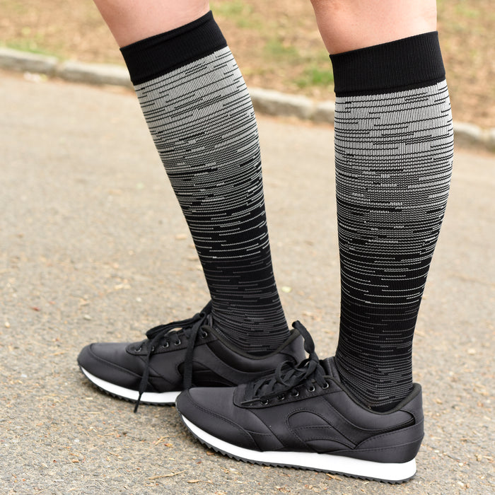 Black and gray ombre compression socks worn with black athletic shoes on a paved surface.