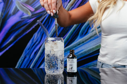 A person holding a dropper above a glass of water with an open bottle of Night Water Infusion Drops | Pure Inventions on a reflective surface, with an abstract blue and purple background.