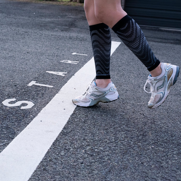 Compression calf sleeves in black with a wave pattern worn by a person in white athletic shoes on a starting line.