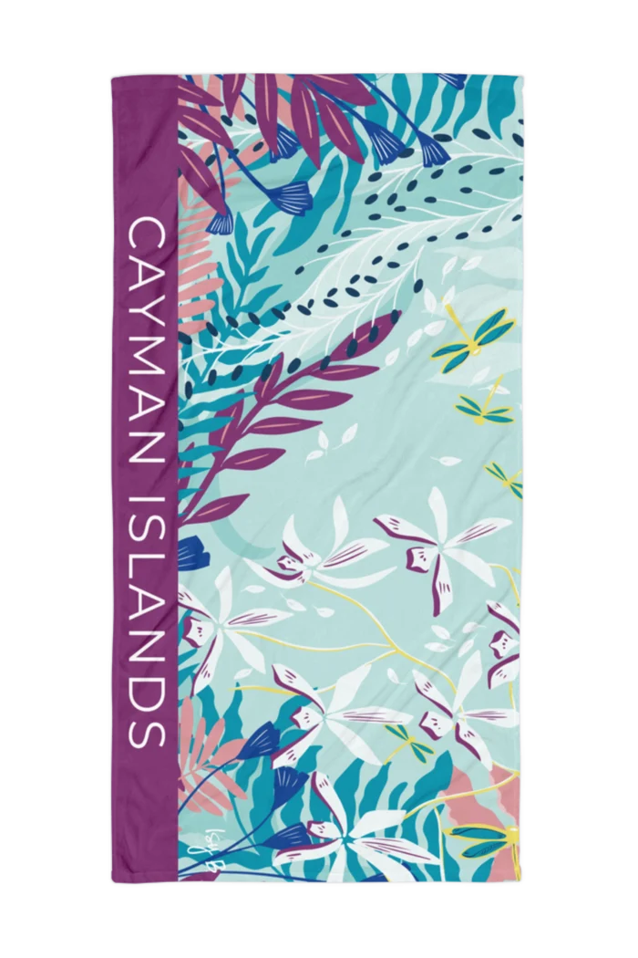 A rectangular beach towel featuring a tropical floral print with the words 'CAYMAN ISLANDS' printed vertically in white on a purple side panel, set against a plain white background.