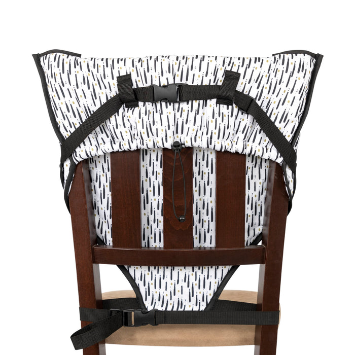 The Portable Toddler Travel Chair features a white patterned fabric with black and yellow dashes, attaches to wooden chairs with black straps, and includes a five-point safety harness for secure child seating.