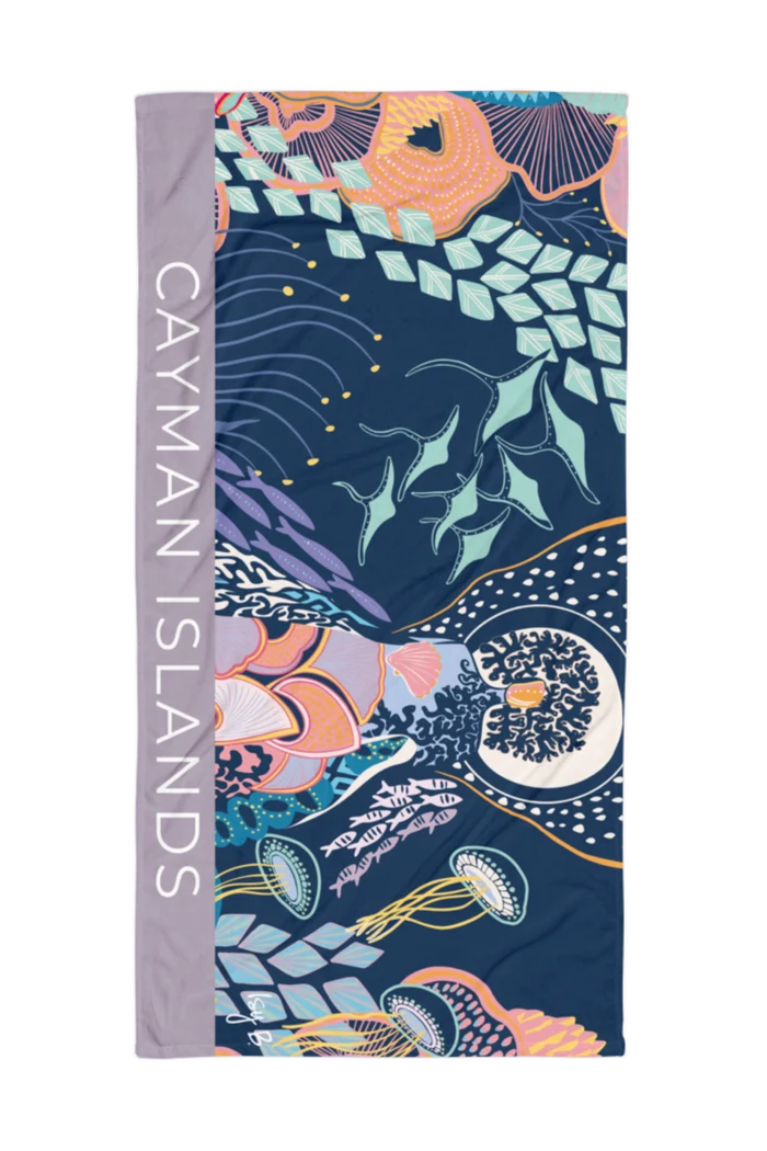 A rectangular beach towel featuring a colorful underwater design with sea creatures on a dark navy background, with a lavender-gray border on the left side printed with the text 'CAYMAN ISLANDS' in white.