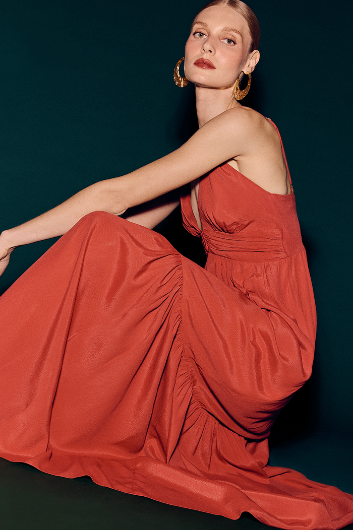 Model wearing long terracotta orange flowing dress with hoop earrings on dark teal background