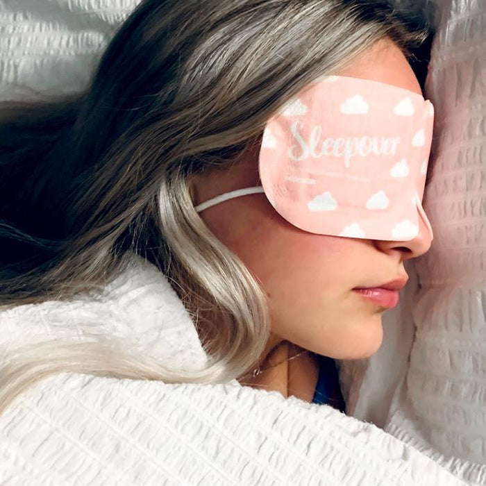 Woman resting in bed wearing a pink Sleepover Self Warming Steam Mask | Popmask with white cloud patterns, promoting relaxation and skincare wellness.