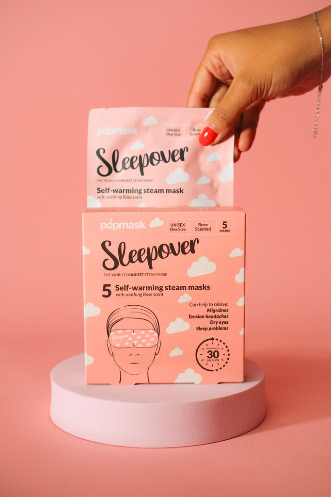 Hand holding a pink Sleepover Self Warming Steam Mask | Popmask packet above a matching box on a cylindrical pedestal against a pink background, highlighting skincare and relaxation benefits.