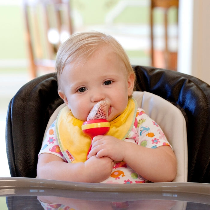 A baby in a high chair chews on the Nuby Nibbler Feeder with a squeeze and twist locking system, wearing a colorful shirt and yellow bib. The feeder uses The Nibbler Feeder Replacement Net - 3 pack for safe enjoyment of whole foods.