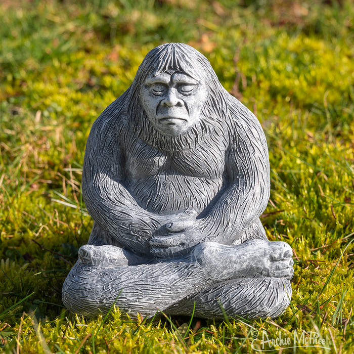 A serene Zen Bigfoot statue, crafted to resemble a stone yeti, sits cross-legged on the grass with its arms resting on its knees and eyes closed, exuding a peaceful aura.